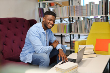 Young man studying in college library, sitting with laptop, surrounded by bookshelves, focused on...