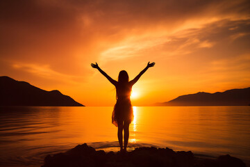 Fototapeta premium Silhouette of a woman with arms raised standing by a serene lake at sunset, surrounded by mountains, embracing the beauty and tranquility of nature
