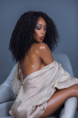 Portrait of serene Black woman with long curly black hair and beautiful makeup sitting by herself on lounge chair inside studio with grey background wearing brown silk dress shirt.