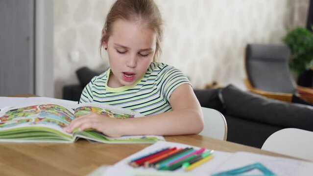 A charming blonde school-age girl is doing her homework while sitting at the kitchen table. She reads aloud and does her homework. The idea of online and home education.