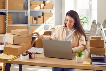 Smiling female small business owner working in warehouse checking stock and inventory sitting at...