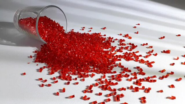 Transparent red polycarbonate masterbatch granules spill from a shot glass over a white background crossed by dramatic lines of light, this polymer is a colorant for products in the plastics industry