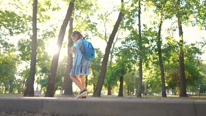 little girl walking along the curb. concept of a happy childhood and loving family. a schoolgirl in a dress and with a blue lifestyle backpack walks along the curb, keeps her balance