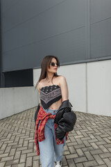 urban beautiful fashionable woman with cool sunglasses in a fashion bandana top and a leather jacket with jeans walks near a modern building