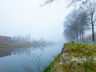 A serene mist envelops a tranquil winter morning near a calm river, reflecting bare trees and fog on its surface
