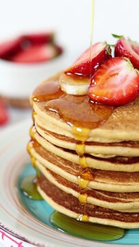 Pouring maple syrup on a stack of pancakes topped with fresh strawberries and bananas for breakfast. Vertical video. Close-up.