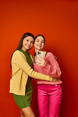 Two pretty teenage girls, brunette and casually dressed, standing side by side in front of a vibrant red wall.