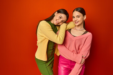 Two pretty brunettes, teenage friends, standing together in front of a vibrant red wall in a studio setting.