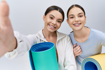 Two pretty and brunette teenage girls, female friends, in sportive attire standing next to each other with yoga mats in a studio on a grey background.