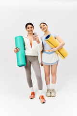 Two pretty teenage girls, one brunette, standing together in sportive attire holding yoga mats on grey studio background.