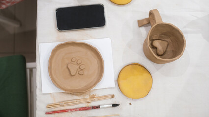Top view of a table in a pottery workshop on which there are products made by hand from clay by student. A plate with the shape of a dog paw and a mug with a heart on the bottom There are tools nearby