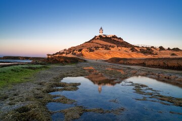 view of the Cabo de la Huerta lighthouse at sunrise with reflections in tidal pools in the foreground