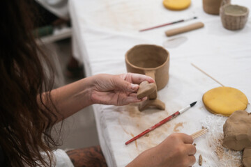 A close-up of the hands of a young woman as she shapes a heart out of clay to decorate a mug....