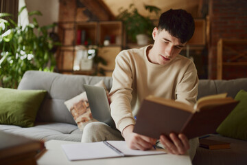 Focused and concerted man, student sitting on sofa at home with modern design and doing homework,...