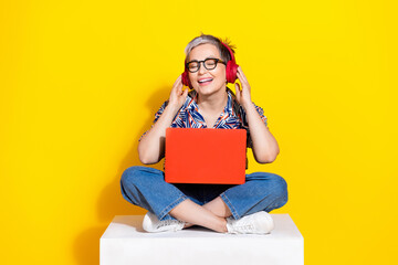 Full length photo of lovely senior lady sit cube netbook headphones dressed stylish colorful garment isolated on yellow color background