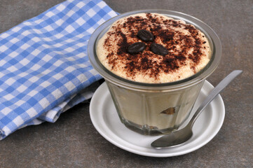 Tiramisu au café dans un ramequin posé sur une soucoupe avec une cuillère et une serviette à carreaux en gros plan sur fond gris