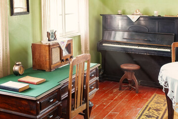 Antique study with desk, books, clock, radio and black piano. The interior is illuminated by...