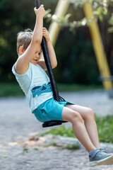 Side view of cute little boy sliding on outdoor rope swing in summer park, blurred background