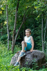 Portrait of a lively cute boy sitting on a stone on a beautiful green background of trees