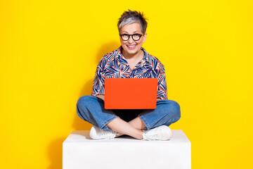 Full length photo of lovely senior lady sit cube hold netbook dressed stylish colorful garment isolated on yellow color background