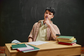 Essence of Learning. Young male student in casual attire sitting at wooden desk with notes and books around, thoughtful face. Blackboard background. Concept of education, studying, homework, youth