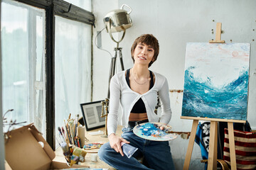 A woman sits in front of a painting, delicately holding a palette.