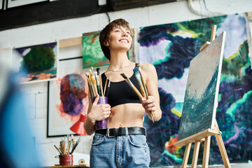 A woman with a paintbrush stands before a painting.