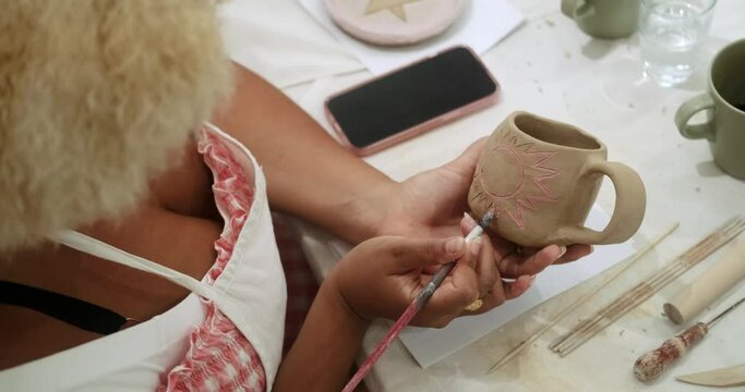 Close-up on the hands of a young African American woman who paints with a brush on a clay mug the sun with special paints. Master class for adults on hand modeling from clay in a bright workshop.