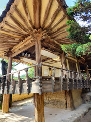 A small wooden building with a balcony and a roof. The roof is made of wood and has a slanted design. The building is surrounded by trees