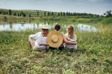 Three young women in casual clothes are sitting by shore of lake or pond and enjoying nature.