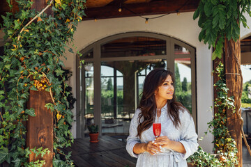 A beautiful brunette girl stands on the terrace of the house and holds a glass of rose wine.