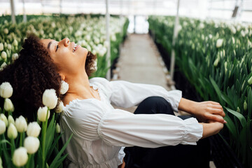 Female portrait of attractive darkskinned girl sitting on path among tulips in greenhouse.
