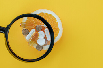 On half of an orange there are different types of tablets and capsules laid out and they are magnified through a magnifying glass. on a yellow background