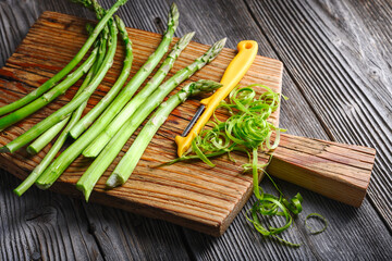 Fresh green asparagus sprouts with yellow vegetable peeler on rustic wooden board close up. Top...