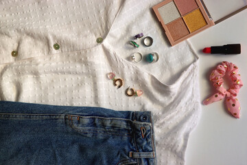 White blouse, denim skirt, colorful rings, earrings and beauty products on white background. Summer outfit in pastel colors. Flat lay.