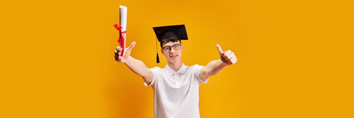 Portrait of young man, student in glasses and graduation cap, with diploma showing positive...