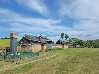 A small house with a fence and a sign in front of it. The house is surrounded by a field and trees