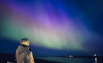 A woman is standing on a pier looking at the sky. The sky is filled with colorful lights of aurora.