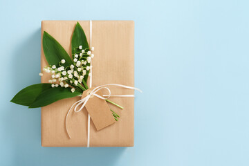 Card with brown gift box with empty rectangular tag and lilies of the valley on blue background....