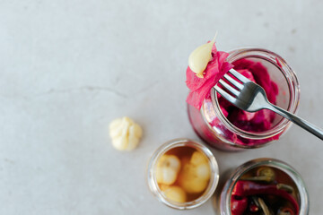 Glass jars with fermented cabbage, garlic and pepper stand on a light stone background. On top of the jar lies a fork with cabbage and garlic