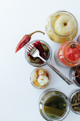 Glass jars with various fermented vegetables stand on a light background. There is a fork with chopped pepper at the top. shot from above