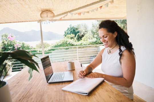 Cheerful woman writing notes during online work on sunny day