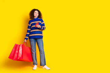 Full length photo of girlish woman wear sweater walk with shopping bags smartphone look empty space isolated on yellow color background