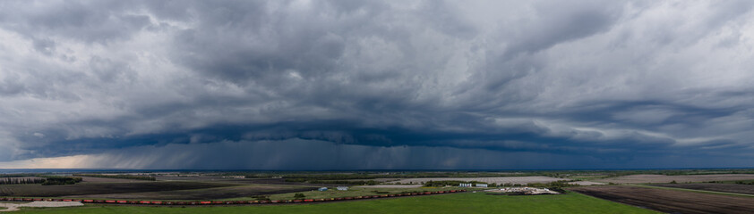 Fototapeta premium Aerial panoramic view of a large blue-gray colored rain and thunder cloud over a flat prairie setting. A train can be seen on a track along the bottom of the image. 