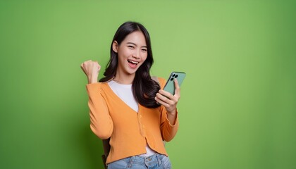  Fun young woman hold in hand use mobile cell phone doing winner gesture clenching fists isolated on green background studio