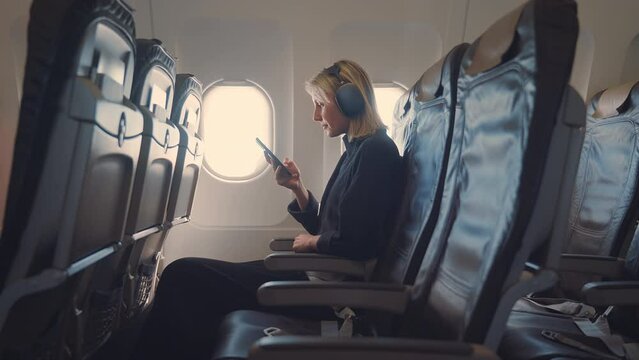 Young blonde woman with headphones using smartphone in airplane. Busy woman sitting near the plane window, texting message on the phone. Working from the sky. 