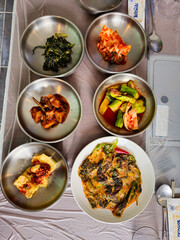 A table with a variety of Asian food, including a plate of broccoli. The table is covered with plastic wrap and has a spoon and a wine glass on it
