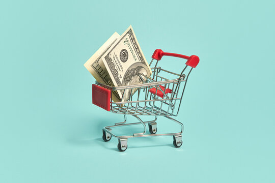 Mini shopping cart with dollar bills on light blue background.