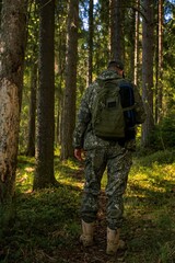 Fototapeta premium View from the back of a brutal man in a military uniform standing in a forest. A ranger with a backpack on a forest path.