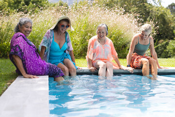 Outdoors, diverse senior female friends sitting by pool, dipping feet in water, copy space